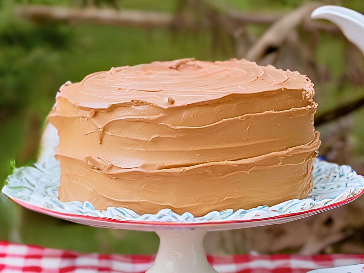 einkorn yellow cake frosted with chocolate buttercream frosting on a white cake stand on a red and white gingham tablecloth