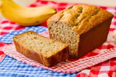 a fresh loaf of banana bread with the end piece cut off sitting on a red gingham table cloth with blue and white checkered napkins beside it and bananas in the background