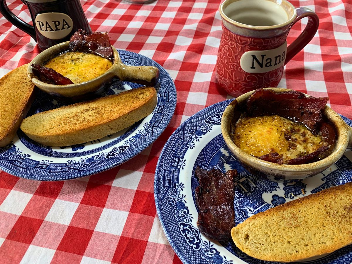 two blue willow plates each with a brown ceramic bowl on it with egg bowl recipe cooked in it with toast and bacon and a black coffee cup that reads Papa and a pink coffee cup that read nana all on a red gingham table cloth