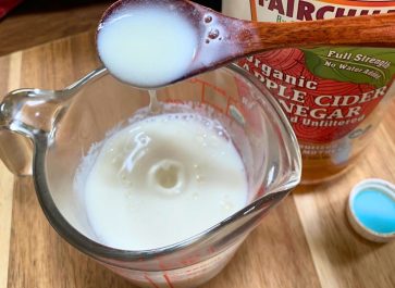 glass measuring cup with homemade buttermilk in it and a wooden spoon is dripping it back into the cup and there is a bottle of apple cider vinegar behind the cup