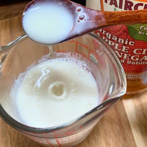 glass measuring cup with homemade buttermilk in it and a wooden spoon is dripping it back into the cup and there is a bottle of apple cider vinegar behind the cup