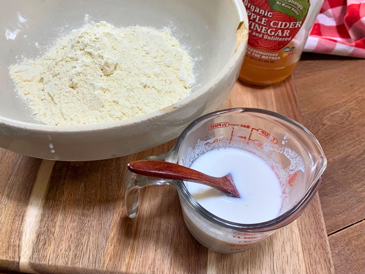 biscuit ingredients in a ceramic mixing bowl with a glass measuring cup beside it with one half cup of homemade buttermilk in it and a bottle of apple cider vinegar behind it